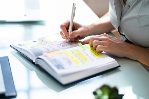 A person writing in an open planner filled with colorful notes and sticky tabs. Their hand holds a pen while the other hand rests on the page. The planner lies on a glass desk with bright natural light in the background.