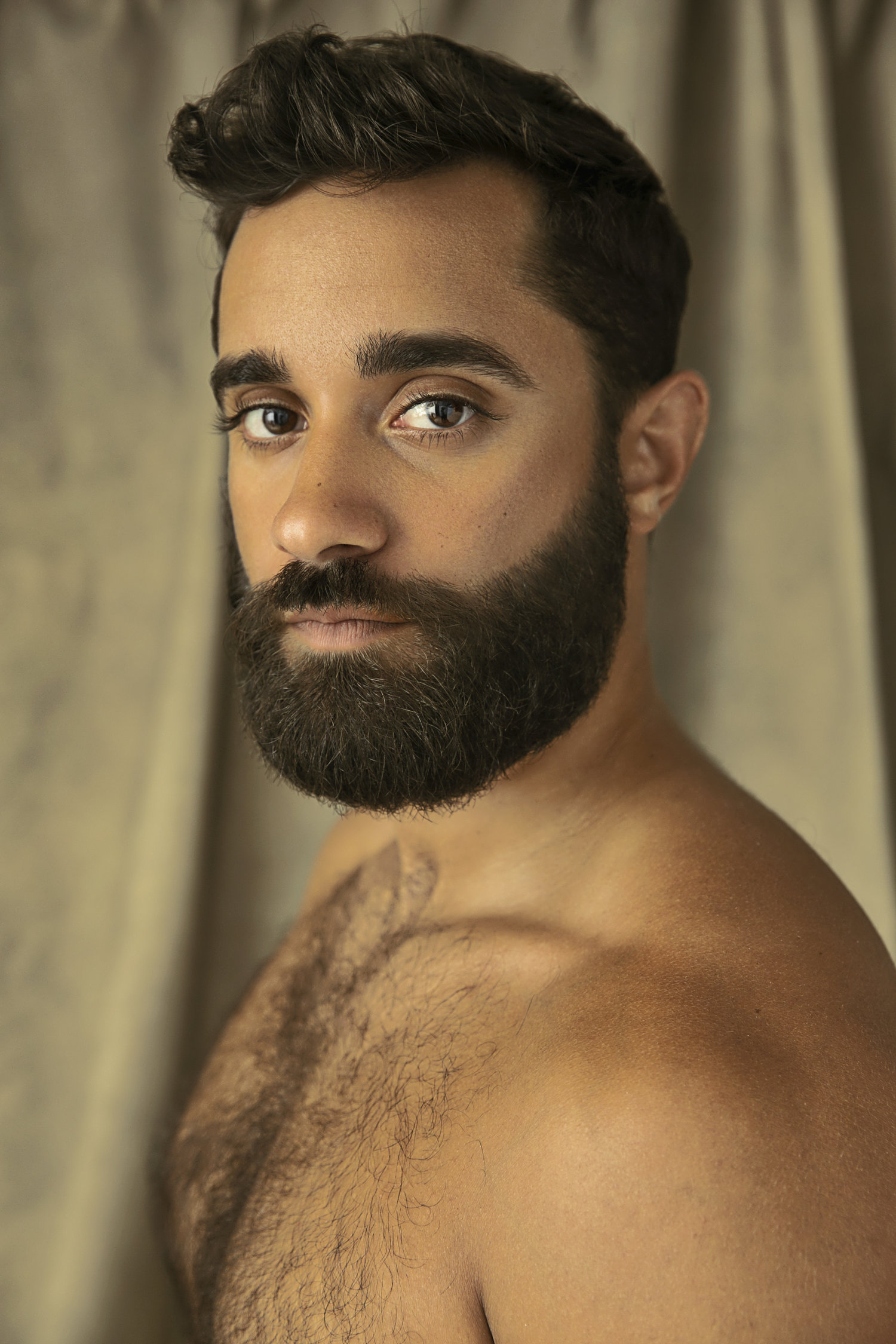 Shirtless close-up of Dr. Julio Torres with a beard and brown eyes, intense gaze against a soft beige background.