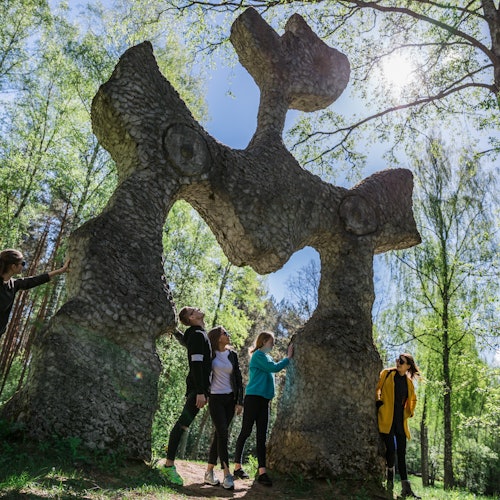 Four people stand beneath a large, abstract stone sculpture in a sunny forest clearing.