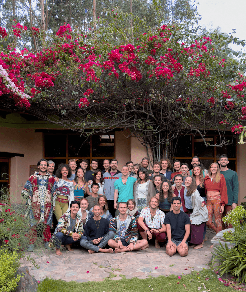 A group yoga teacher students are gathered outdoors, smiling in front of a building with vibrant pink flowers. The setting conveys a sense of community and joy.