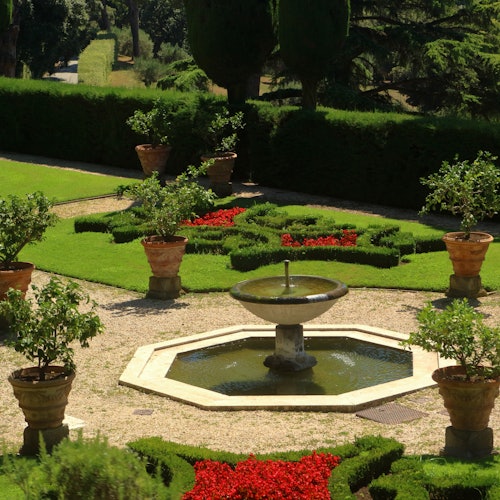一個正式的花園,中央有一個八角形的噴泉,紅色花朵呈幾何圖案,砂石小徑上擺放著盆栽植物。