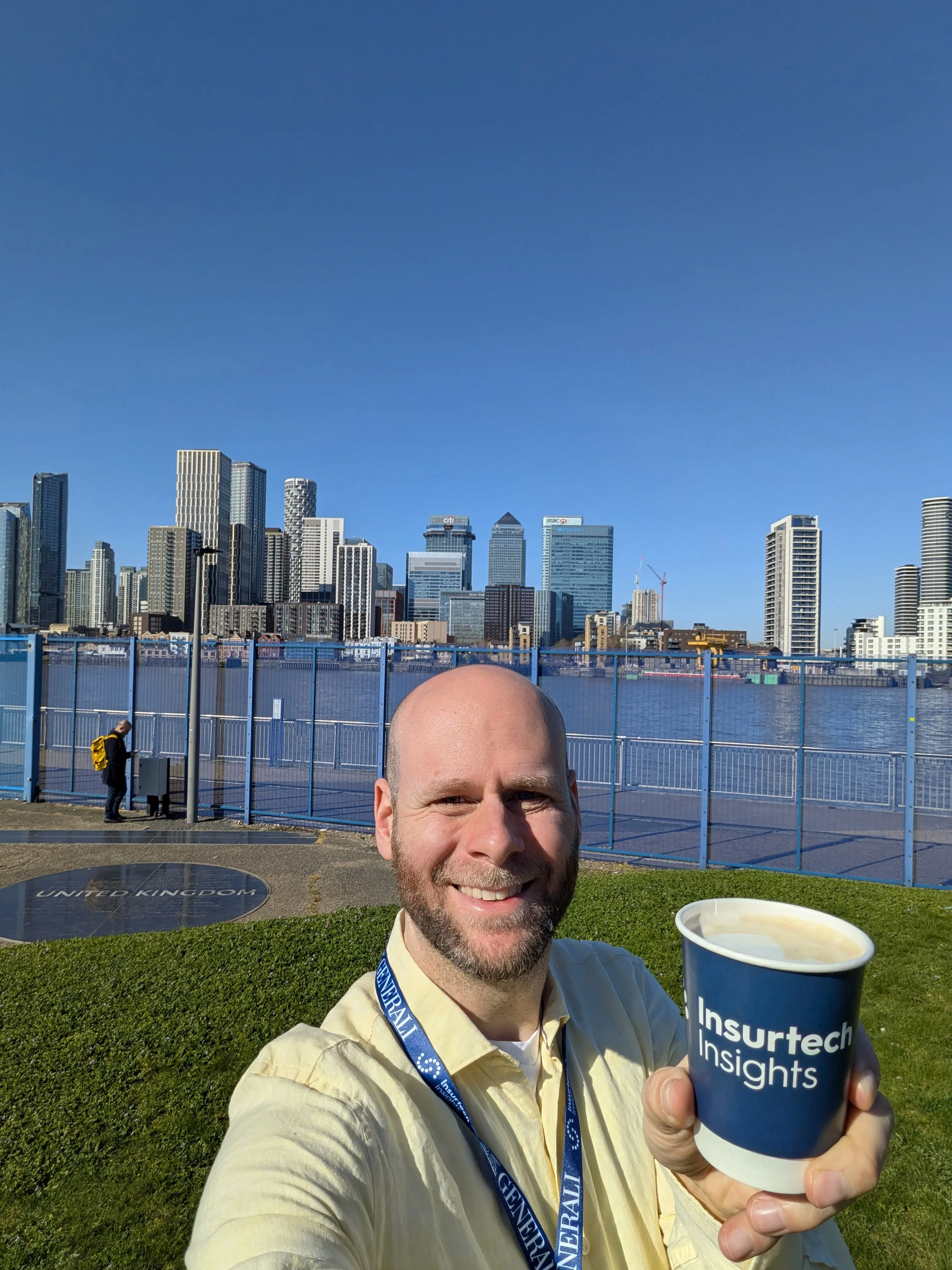 A person stands outdoors, smiling and holding a cup, with a city skyline and blue sky in the background.