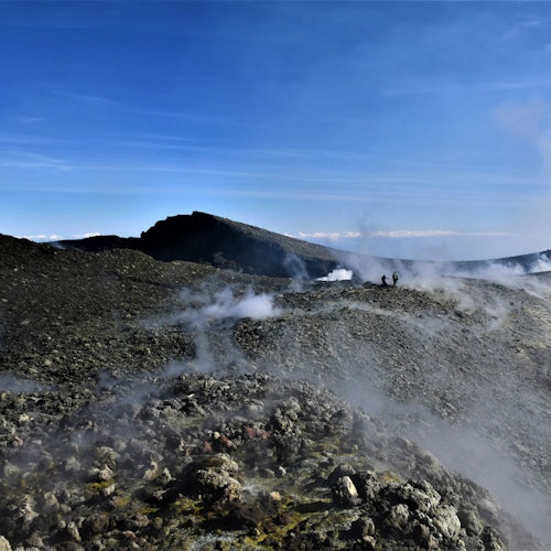 Duas pessoas estão de pé numa paisagem rochosa e fumegante, com montanhas ao longe sob um céu azul claro.