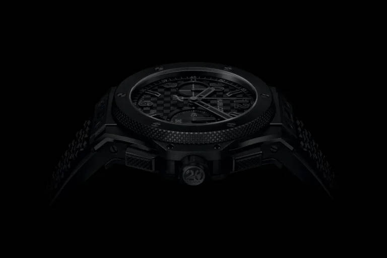 Low-key frontal view of a black luxury wristwatch with textured bezel and patterned dial, emerging from a dark background.