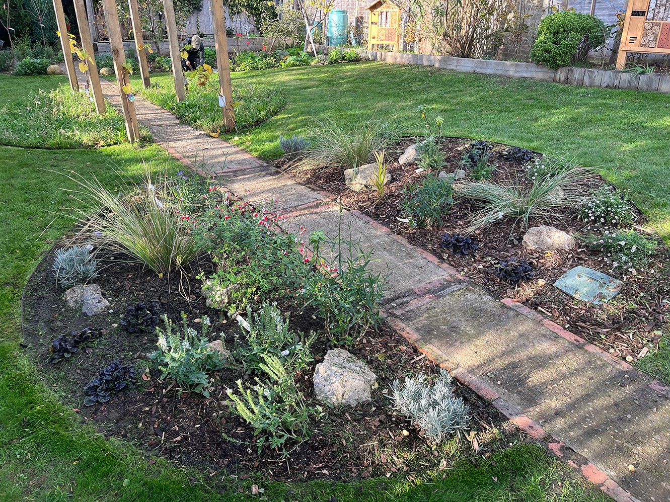 A garden path winding through lush greenery and rounded flower beds with rocks, surrounded by trees.