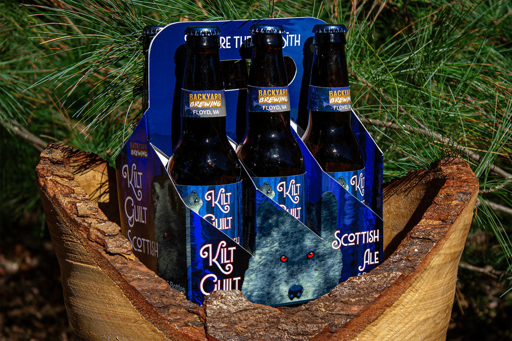 Kilt Guilt Scottish Ale six-pack mockup – moody blue packaging with mystical wolf imagery displayed on tree stump with pine needles