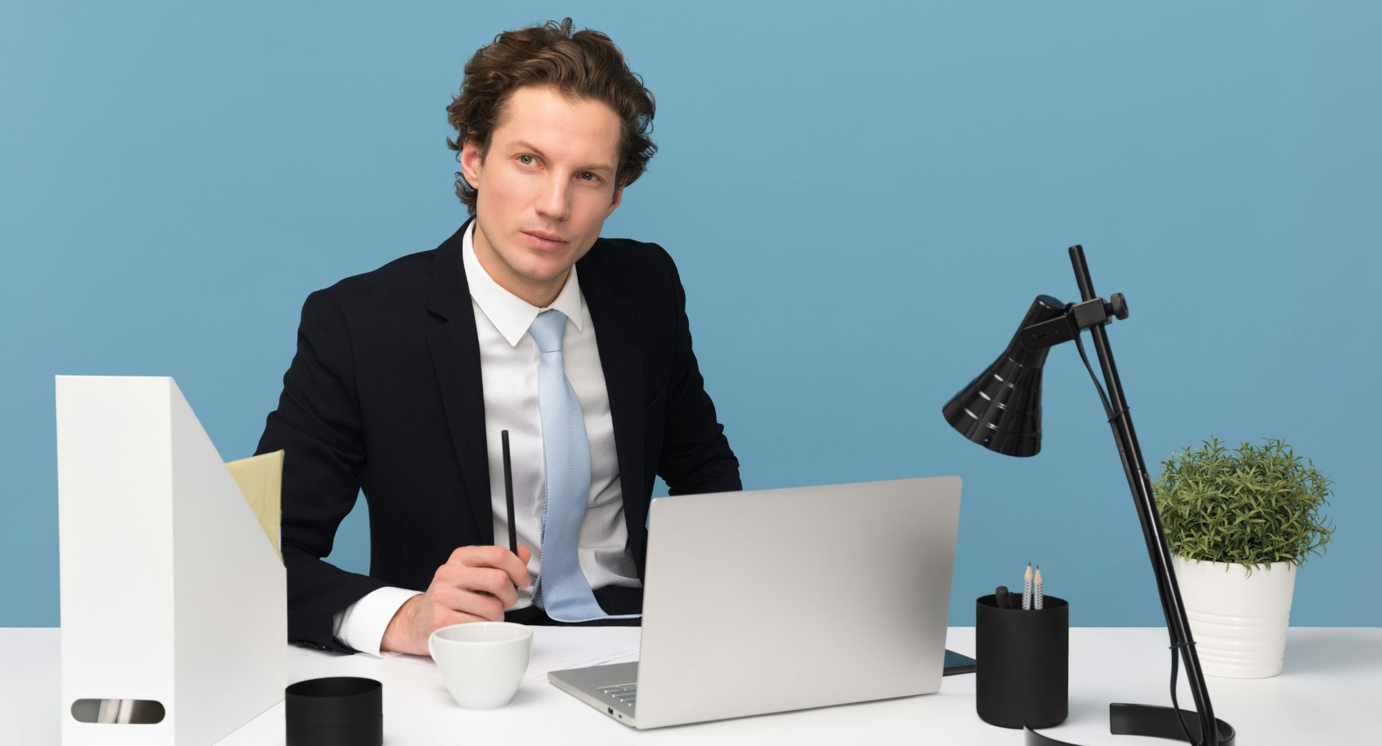 homem de terno e gravata cor azul bebê visto de frente sentado em uma mesa de escritório com notebook e outros objetos, a cor da parede ao fundo é a mesma da gravata