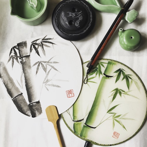 两只带有竹子图案的手持扇子、一支书法毛笔和各种绿色陶瓷物品整齐地摆放在白色的表面上。