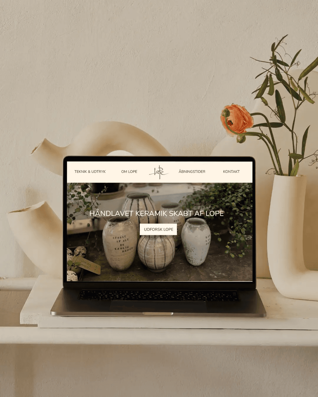 Computer der viser håndlavet keramik hjemmeside, omgivet af vaser og blomster.