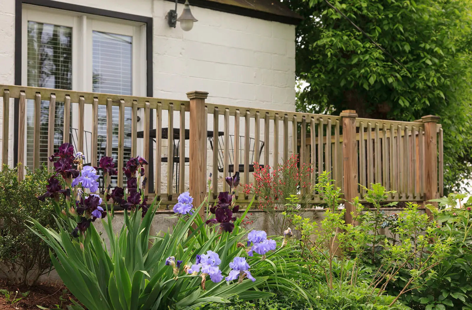 A wooden deck with colorful flowers in front of a house, surrounded by greenery and a wooden fence.
