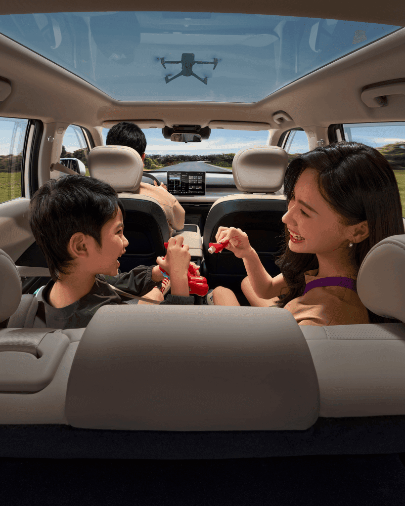 A woman and a child sitting in the backseat of a car, smiling and playing with red toys, while a man is driving in the front. The ceiling features a panoramic view of the sky and a drone is visible above.