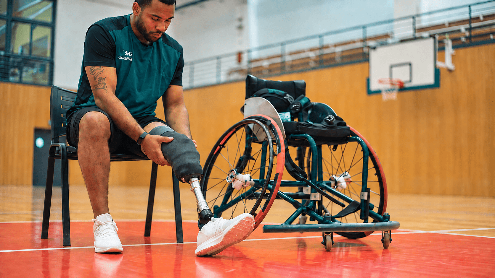 A prosthetic user adjusting a leg prosthesis beside an adaptive wheelchair in a gym.