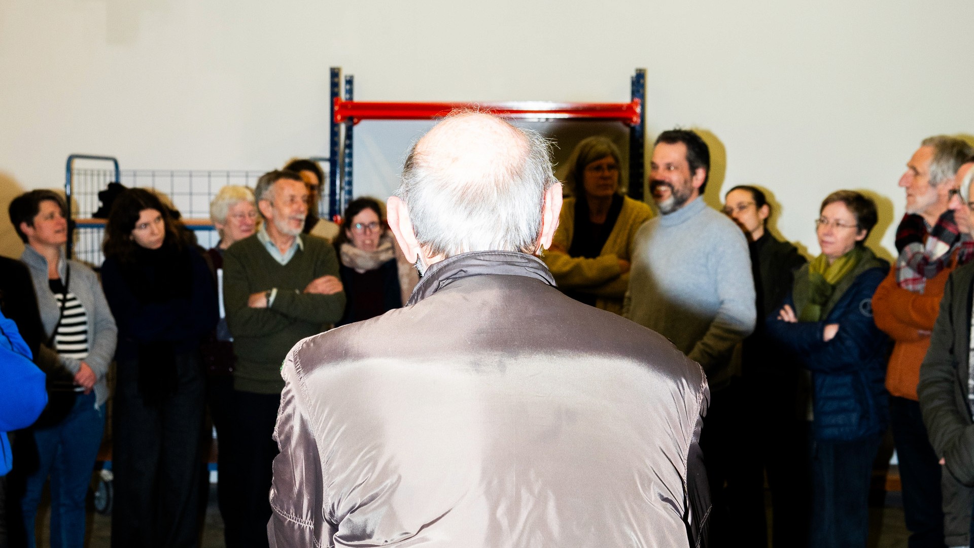 De foto toont een groep mensen die in een halve cirkel luisteren naar iemand die het woord neemt. Het middelpunt van de foto is de rug van een oudere man.