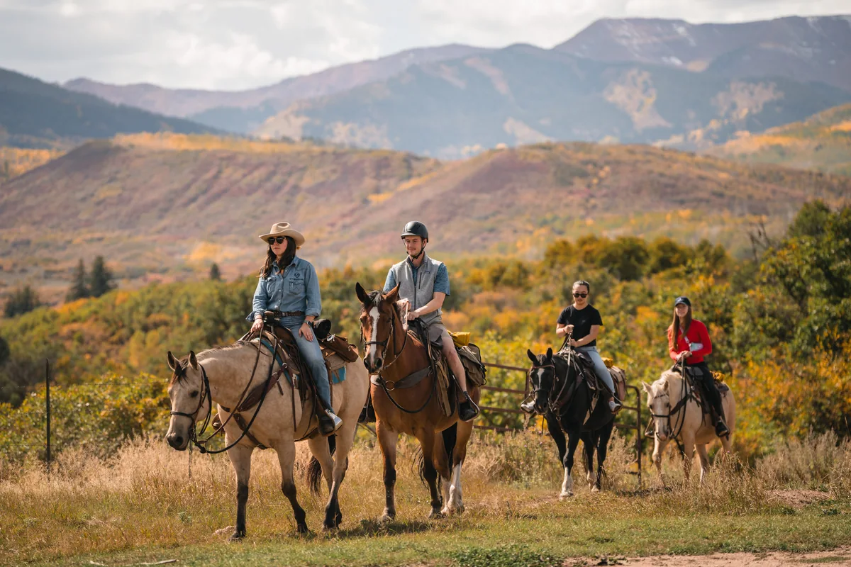 Four horseback riders cross a grassy meadow with autumn hills and mountains behind them.