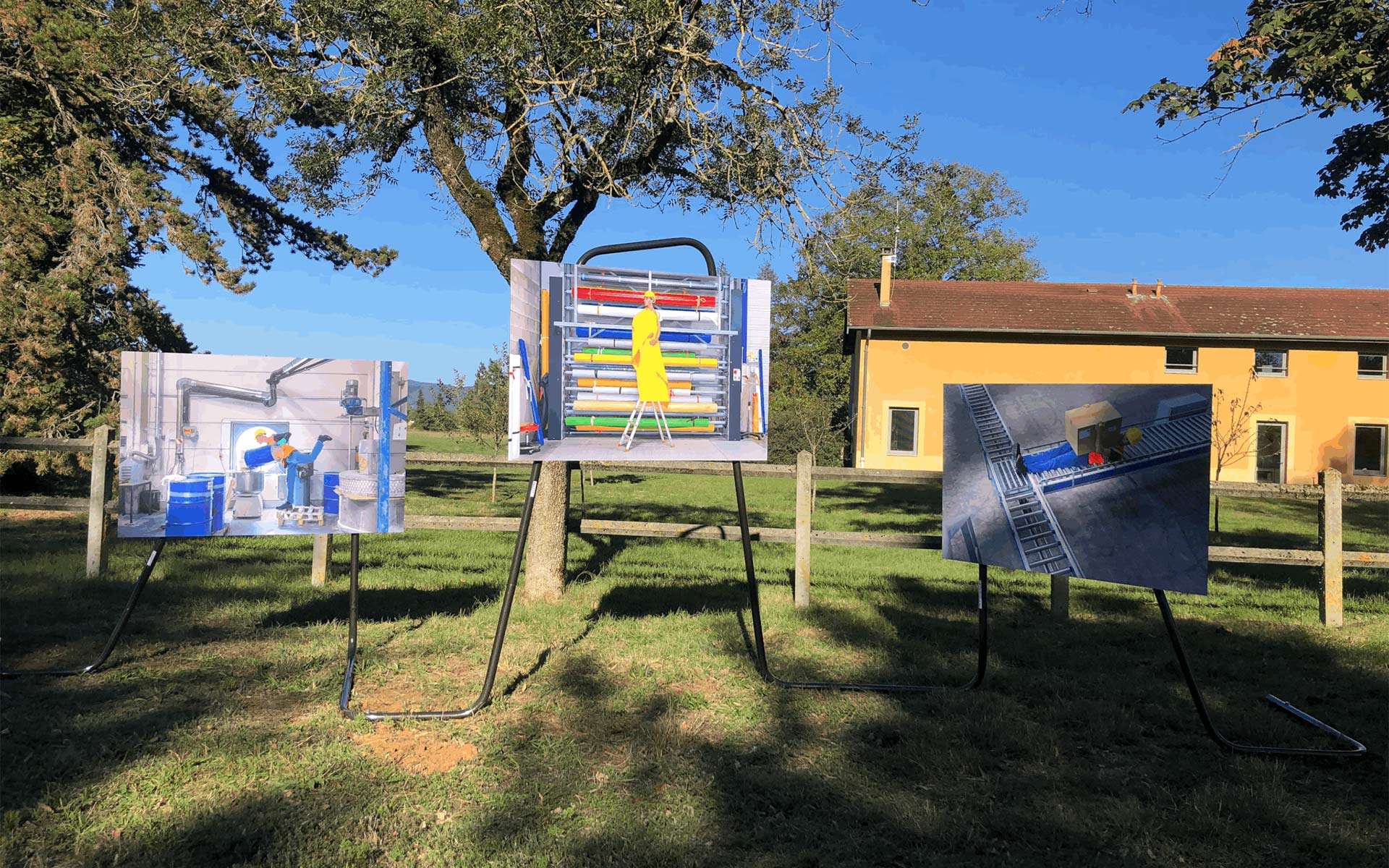 Trois grands tirages photographiques sont installés en extérieur dans un espace verdoyant, sur des supports métalliques indépendants. Ces photos présentent des scènes de travail industriel : à gauche, une personne manipulant des barils bleus ; au centre, une personne en tenue jaune sur un escabeau devant un rayonnage de bobines colorées ; à droite, une personne sur un tapis roulant industriel avec un colis. En arrière-plan, on aperçoit une clôture en bois, un grand arbre et un bâtiment jaune sous un ciel bleu dégagé.