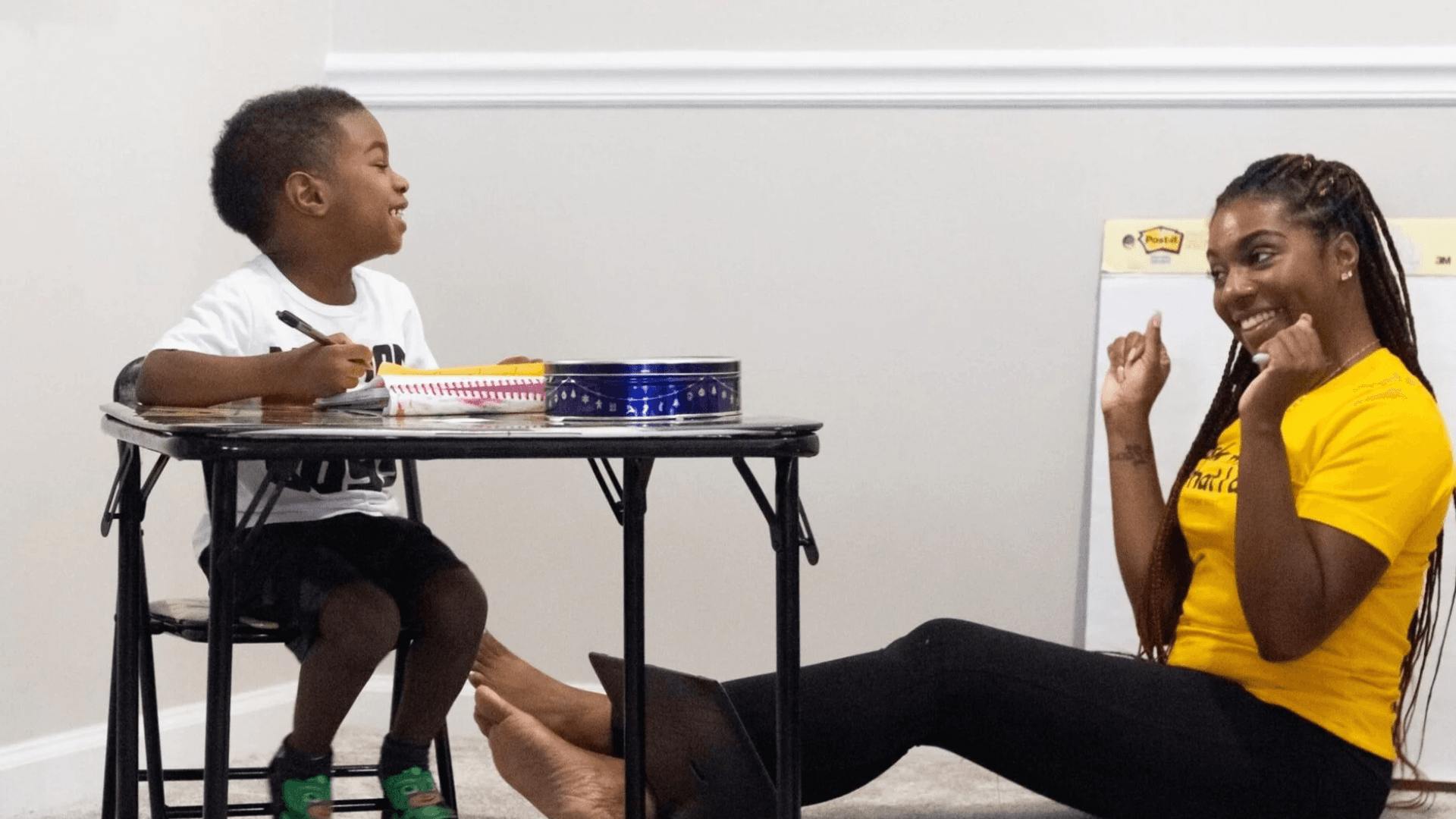 Adult woman sitting on the floor smiling and encouraging a young boy who is seated at a desk drawing and writing in a notebook.