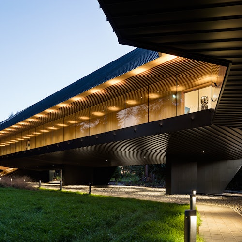 一棟現代建築，擁有大型玻璃窗和外部燈光，懸空在一片綠草地上，旁邊有小徑。