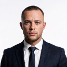 Portrait of a woman with straight long hair, in a dark t-shirt, set against a neutral backdrop.
