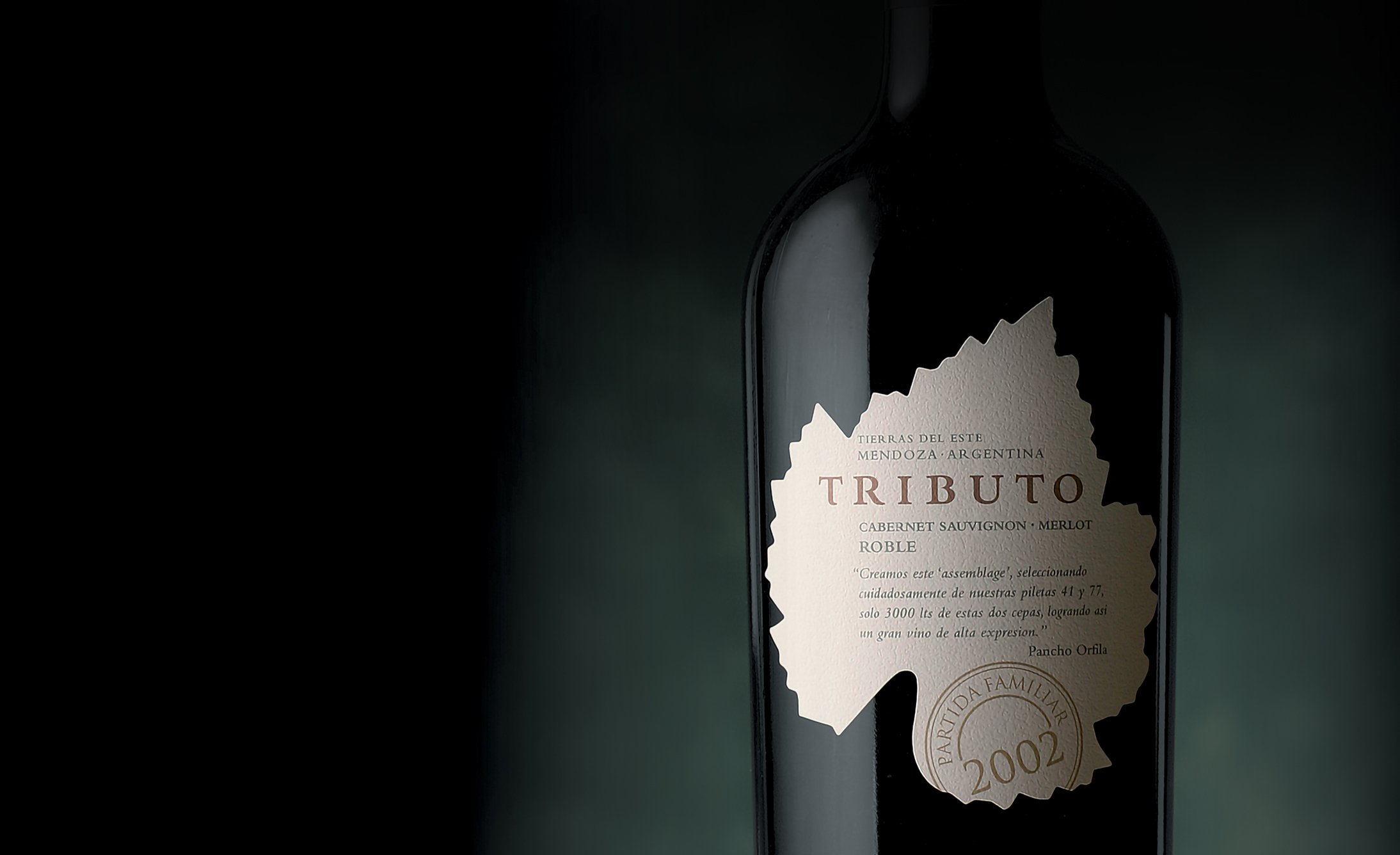 A dark wine bottle with a beige label stands against a muted green background. The label reads "Tributo" and features vintage details, suggesting elegance.
