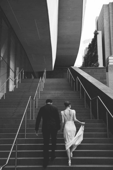 Contemporary wedding portrait in black and white, showing a bride and groom ascending stairs in an urban setting, emphasizing style and simplicity.