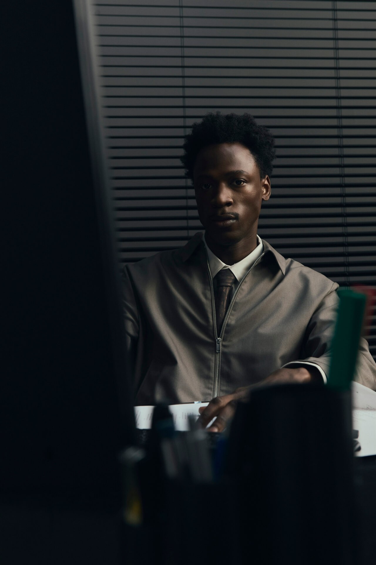 A man in a grey shirt sits in a dim office, gazing seriously at a computer screen. Blinds and office supplies create a focused and professional atmosphere.