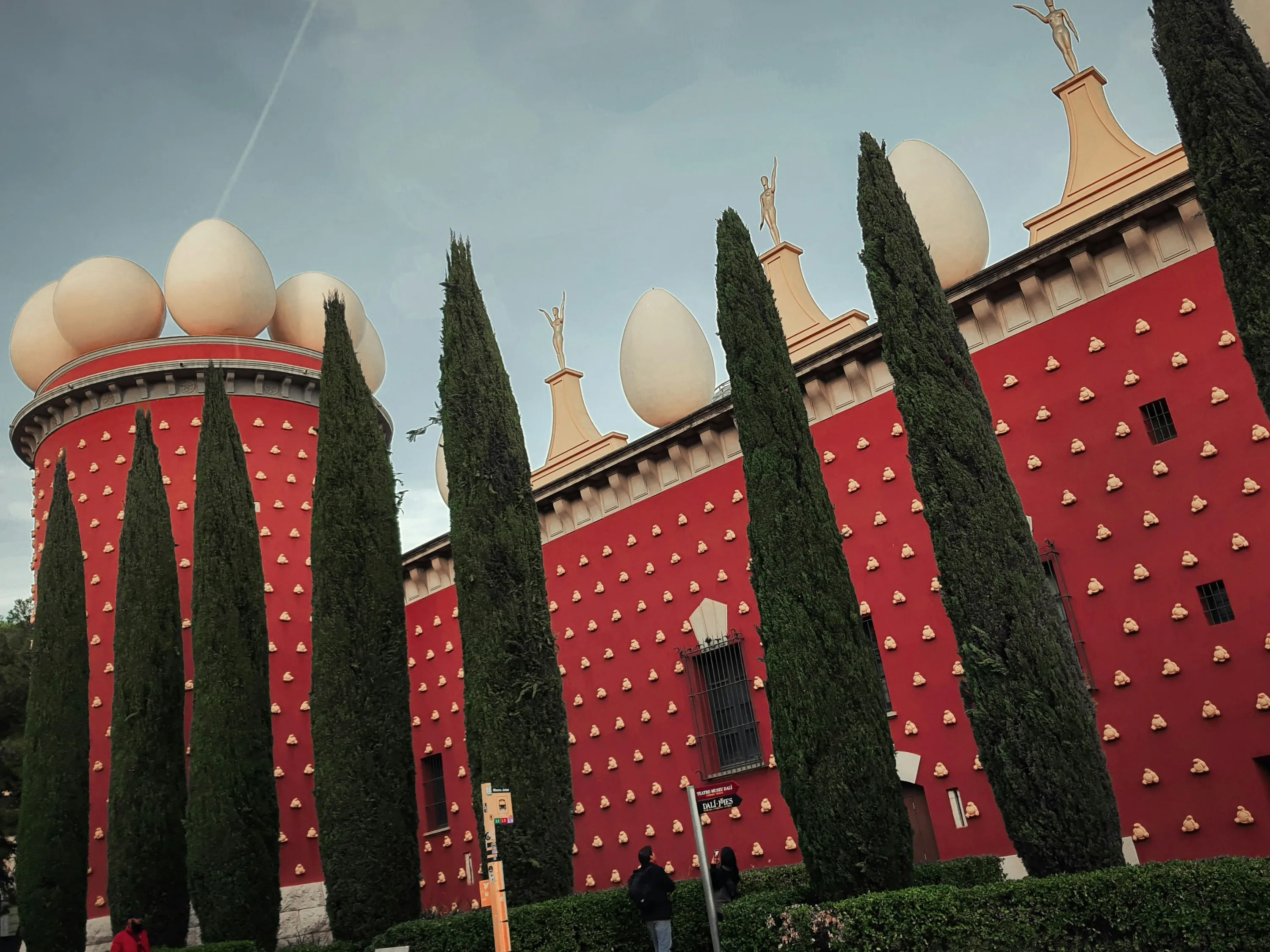 Red facade of the Dali Theatre Museum topped with large egg statues, with green trees at the forefront.