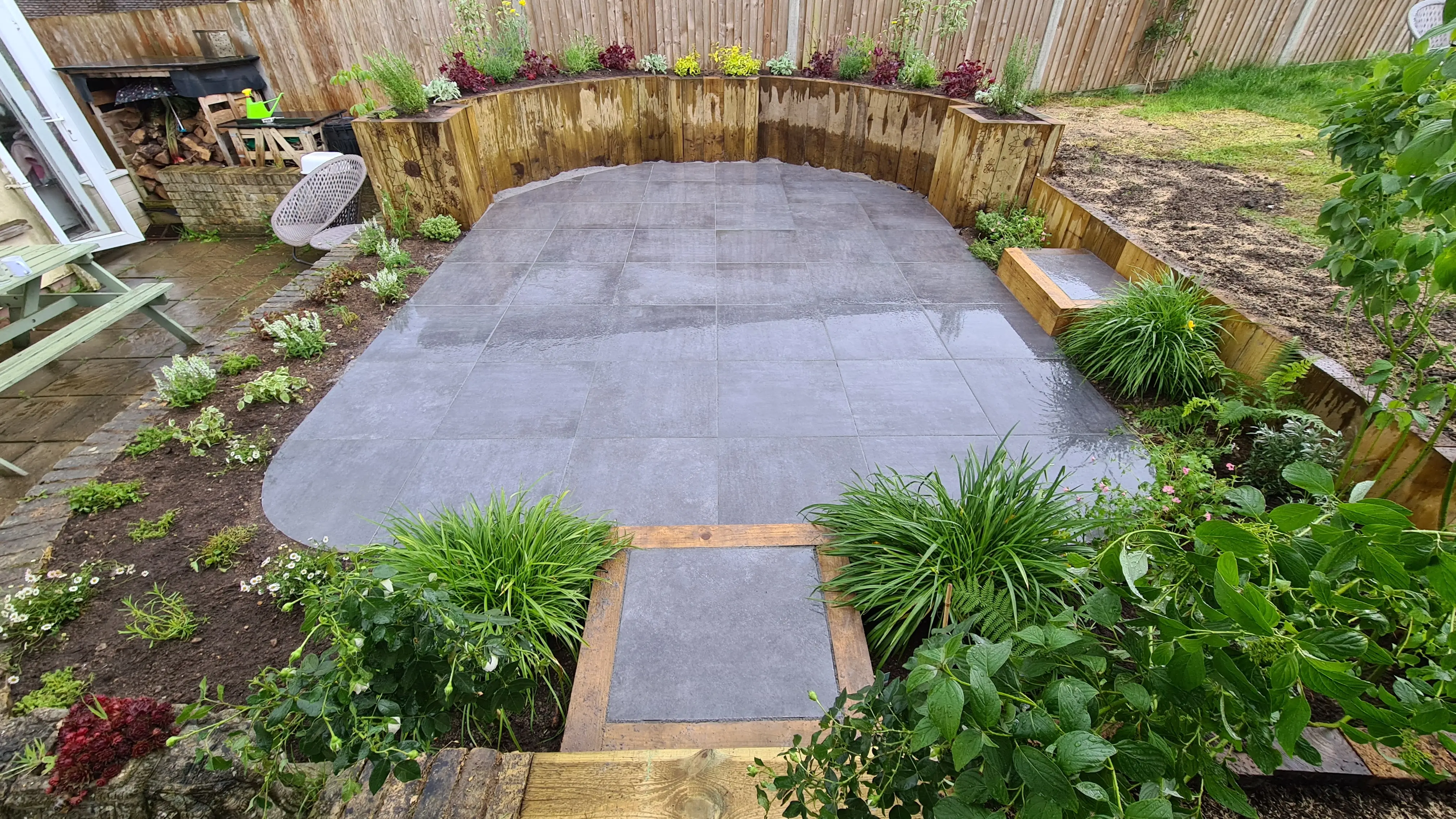 A landscaped backyard with a stone pathway leading to a patio area surrounded by greenery and decorative stones.