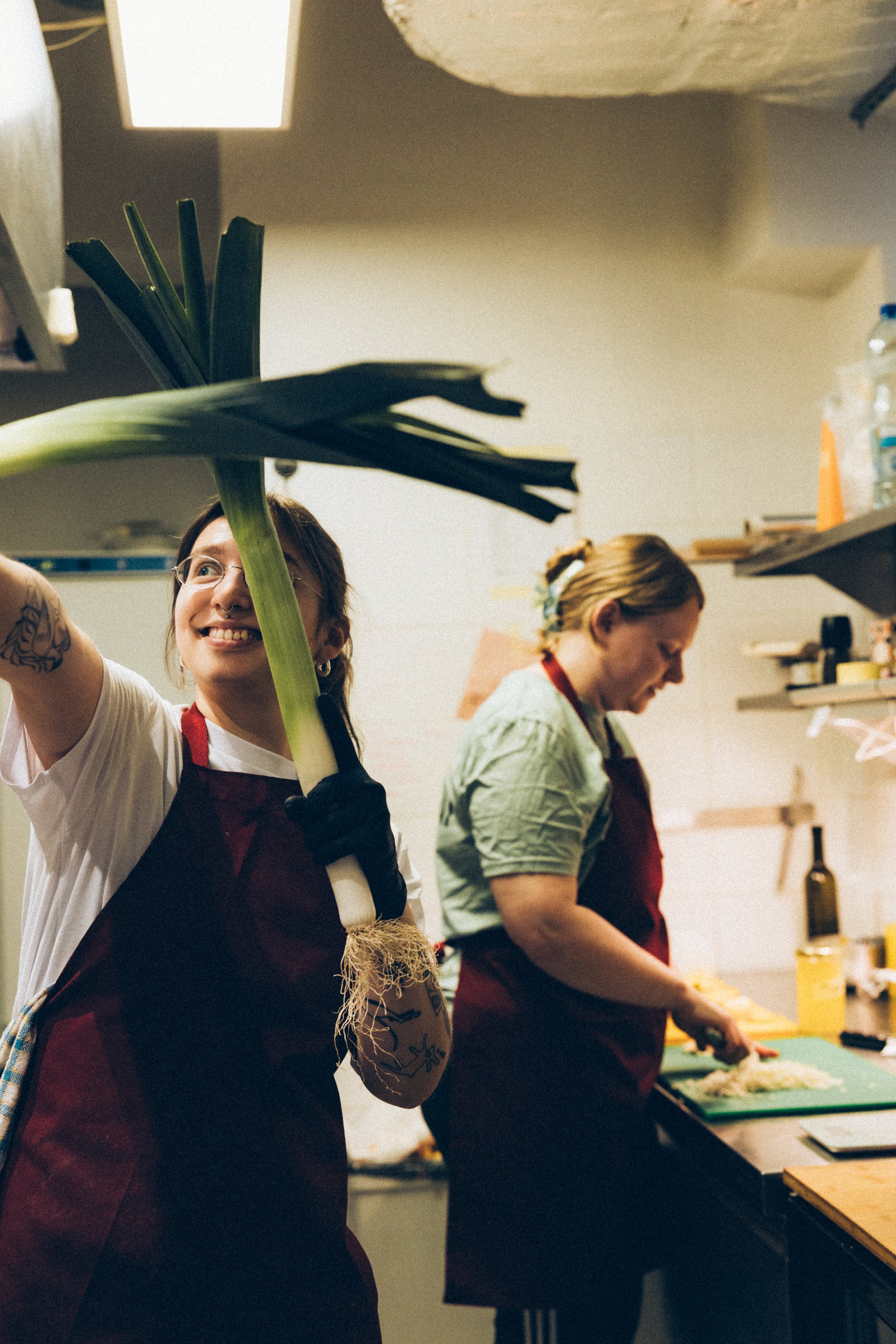 Two workers in aprons are busy in a kitchen, one holding a large utensil while the other prepares food.