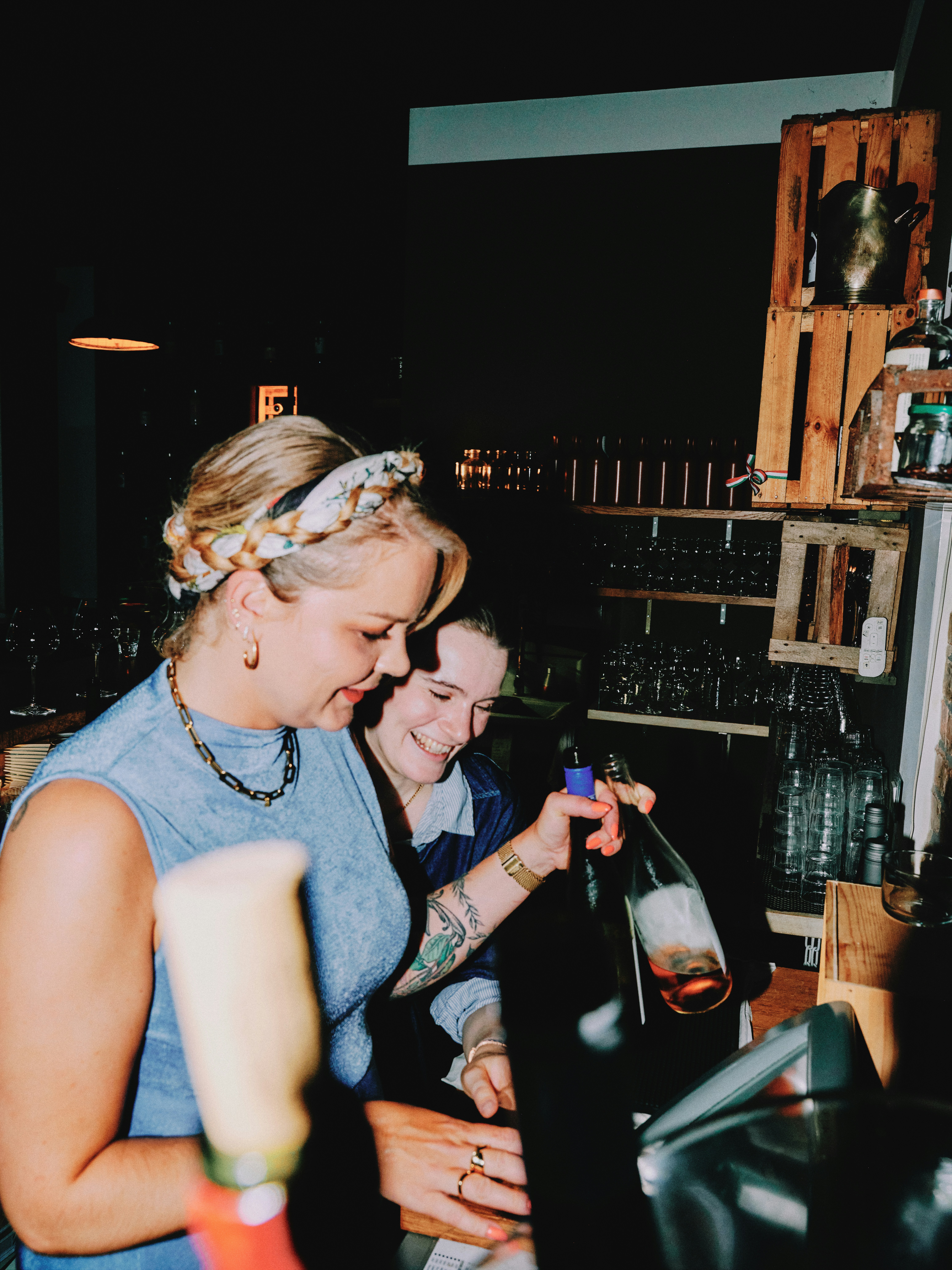 Candid shot of two female wine bar staff laughing together behind the bar counter, wooden crate shelving and glassware visible in the dark background