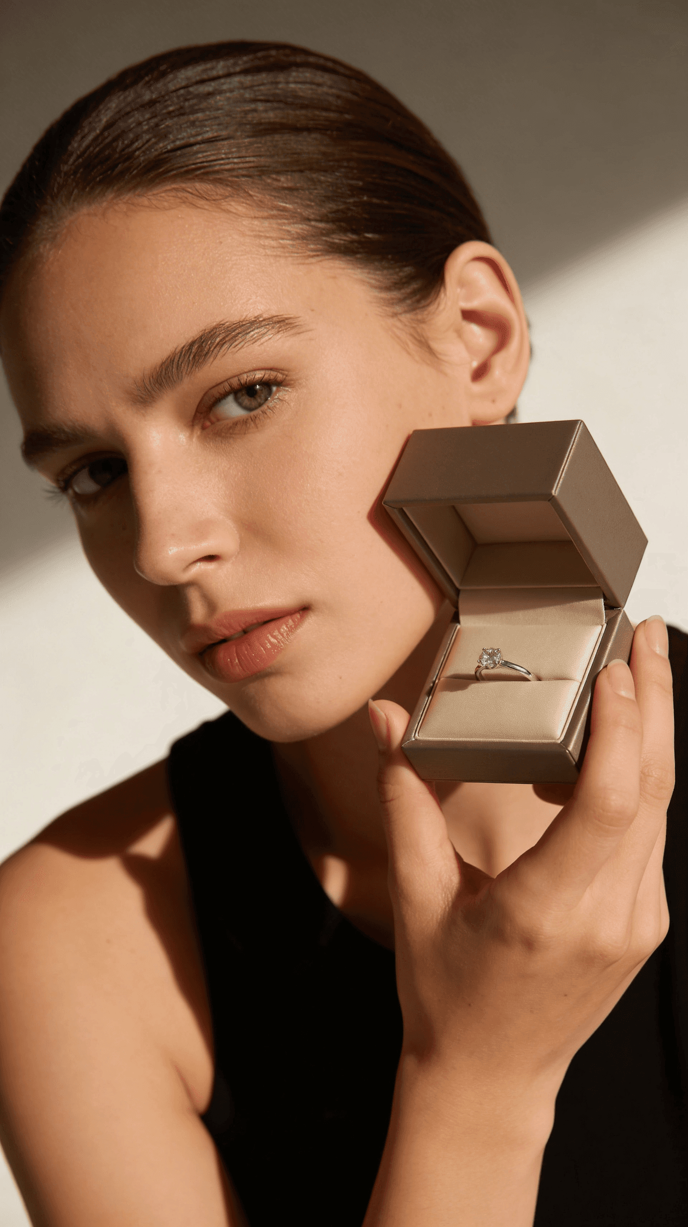 A woman holding an open jewelry box with a diamond ring, set against a neutral background with soft lighting.
