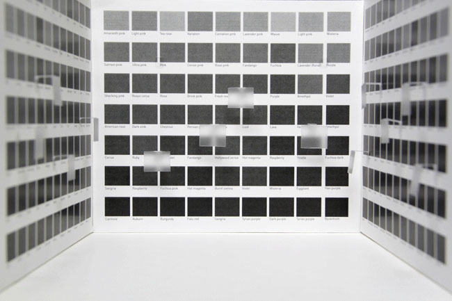 The image shows a white rectangular box divided into two sections by vertical walls, each containing multiple square images arranged in rows and columns. The images are grayscale and appear to be printed on labels or cards, with some labels partially obscured by translucent sheets. The box is positioned on a white surface, and the lighting is even and bright, casting no strong shadows. The images within the box are mostly dark gray or black, with some lighter gray or white areas. No text or labels are visible on the images themselves.