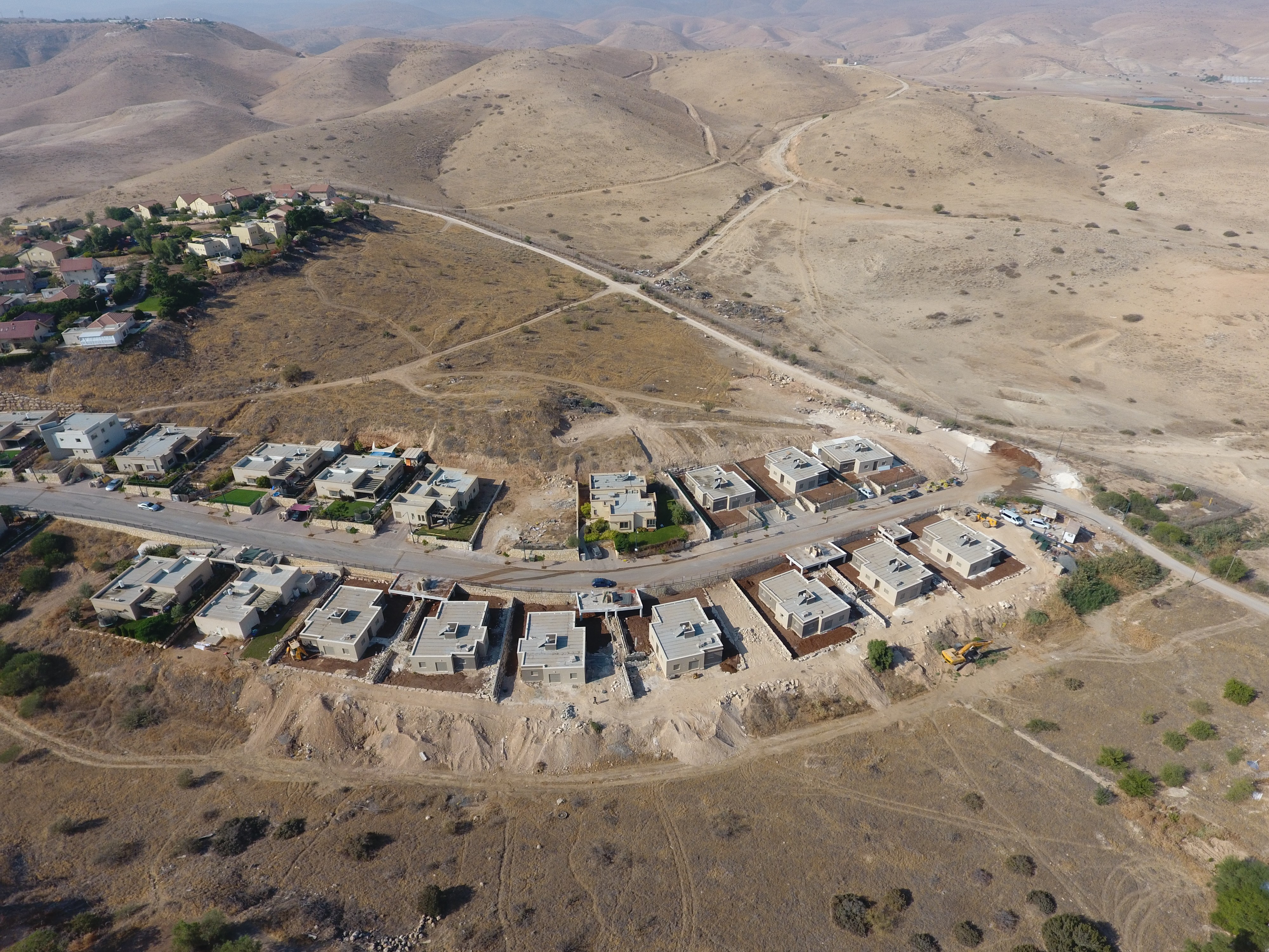 Aerial view of a rural desert landscape featuring a crescent of modern housing with flat roofs, interconnected by a single road, surrounded by vast, barren hills under a clear sky.