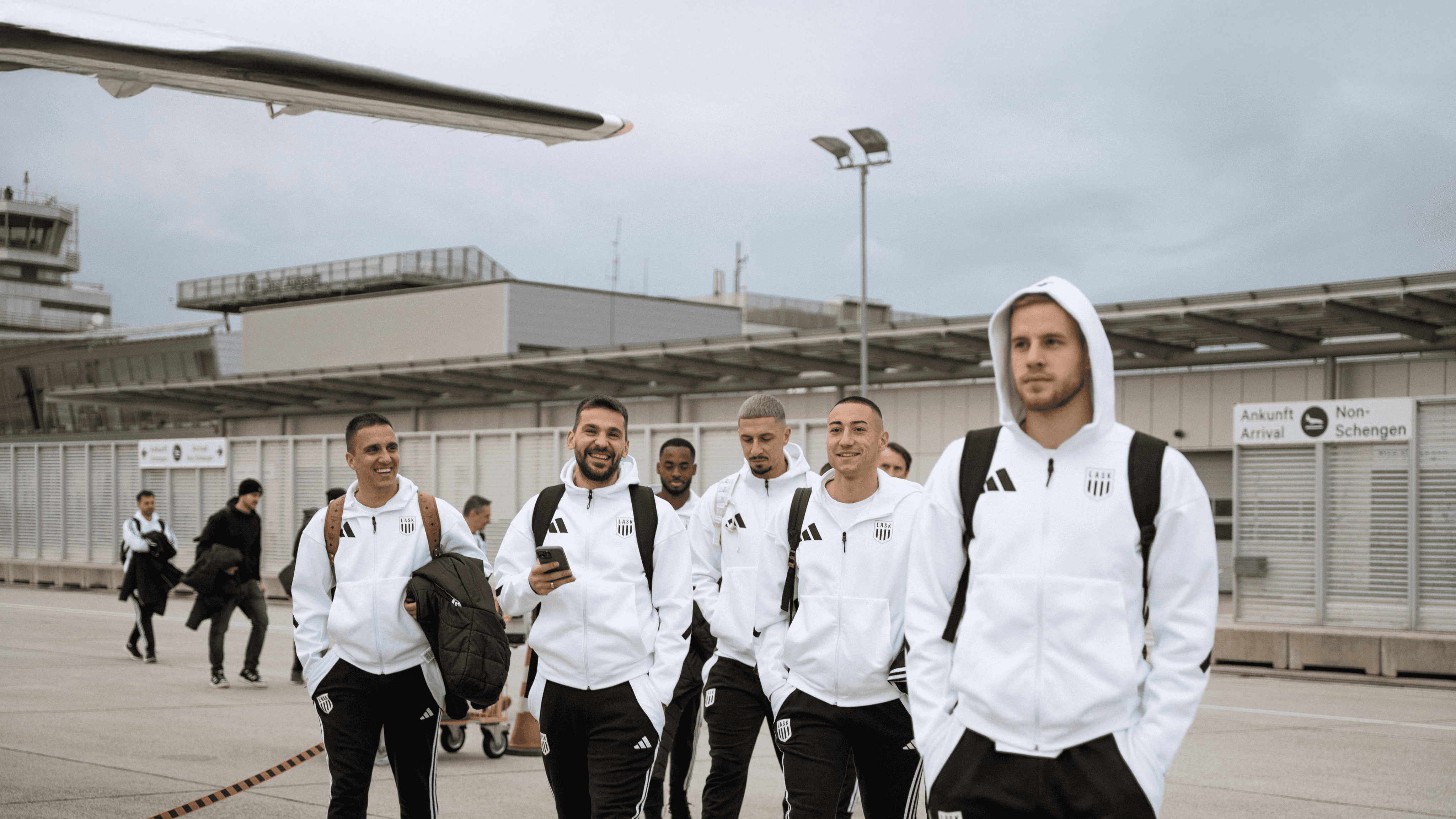 Fußballmannschaft geht vom Flugzeug. Spieler tragen weiße Trainingsjacken und dunkle Hosen. Flughafen im Hintergrund.