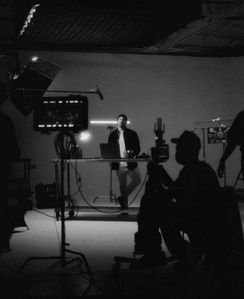 A dimly lit scene with a person standing, surrounded by silhouetted figures and film equipment.