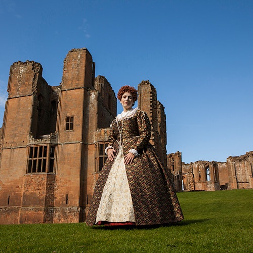 一个穿着古装的人站在一个古老、部分破损的砖建筑前，建筑坐落在青草如茵的草坪上，头顶是晴朗的天空。