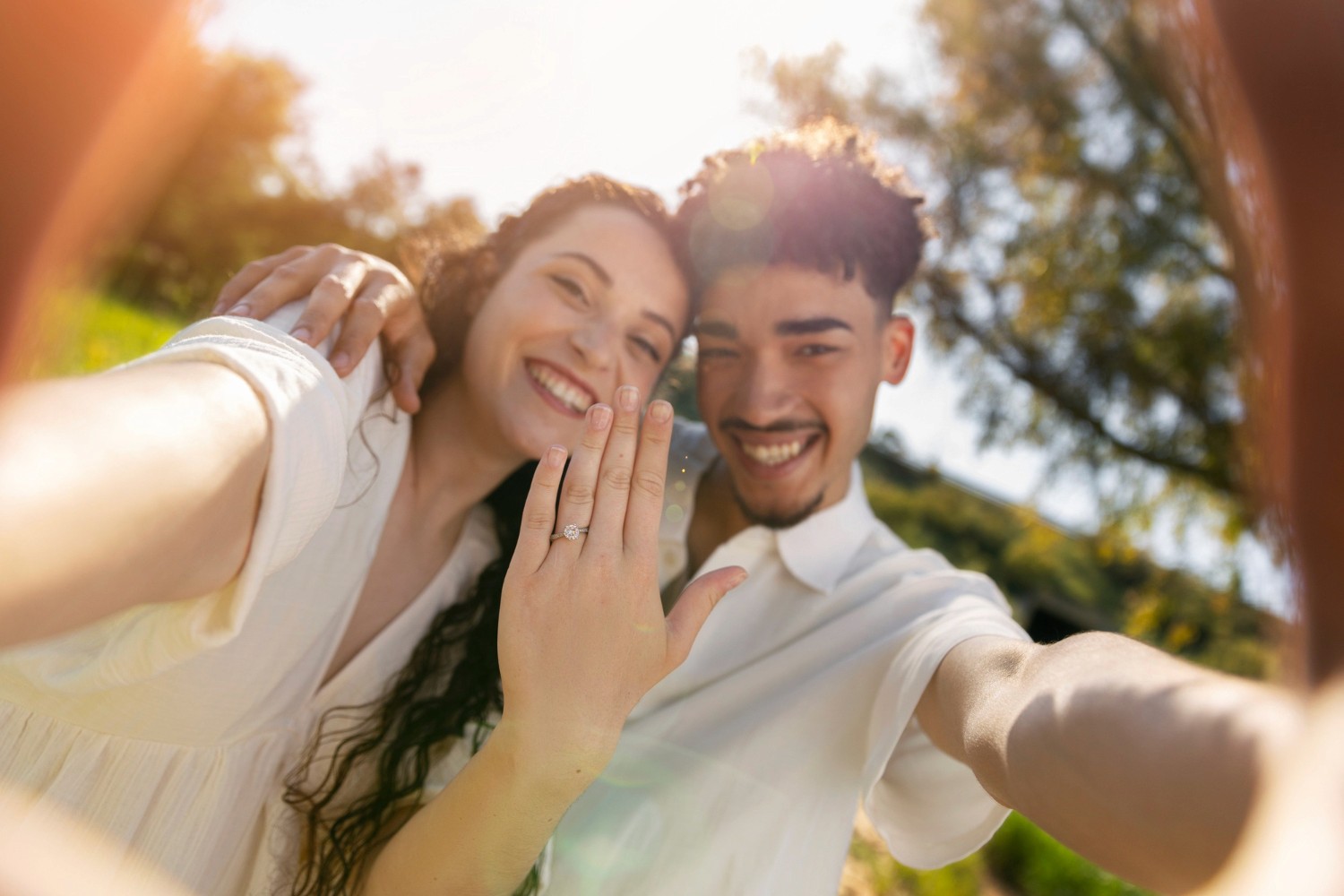 A smiling couple takes a sunny outdoor selfie; the woman shows an engagement ring on her finger.