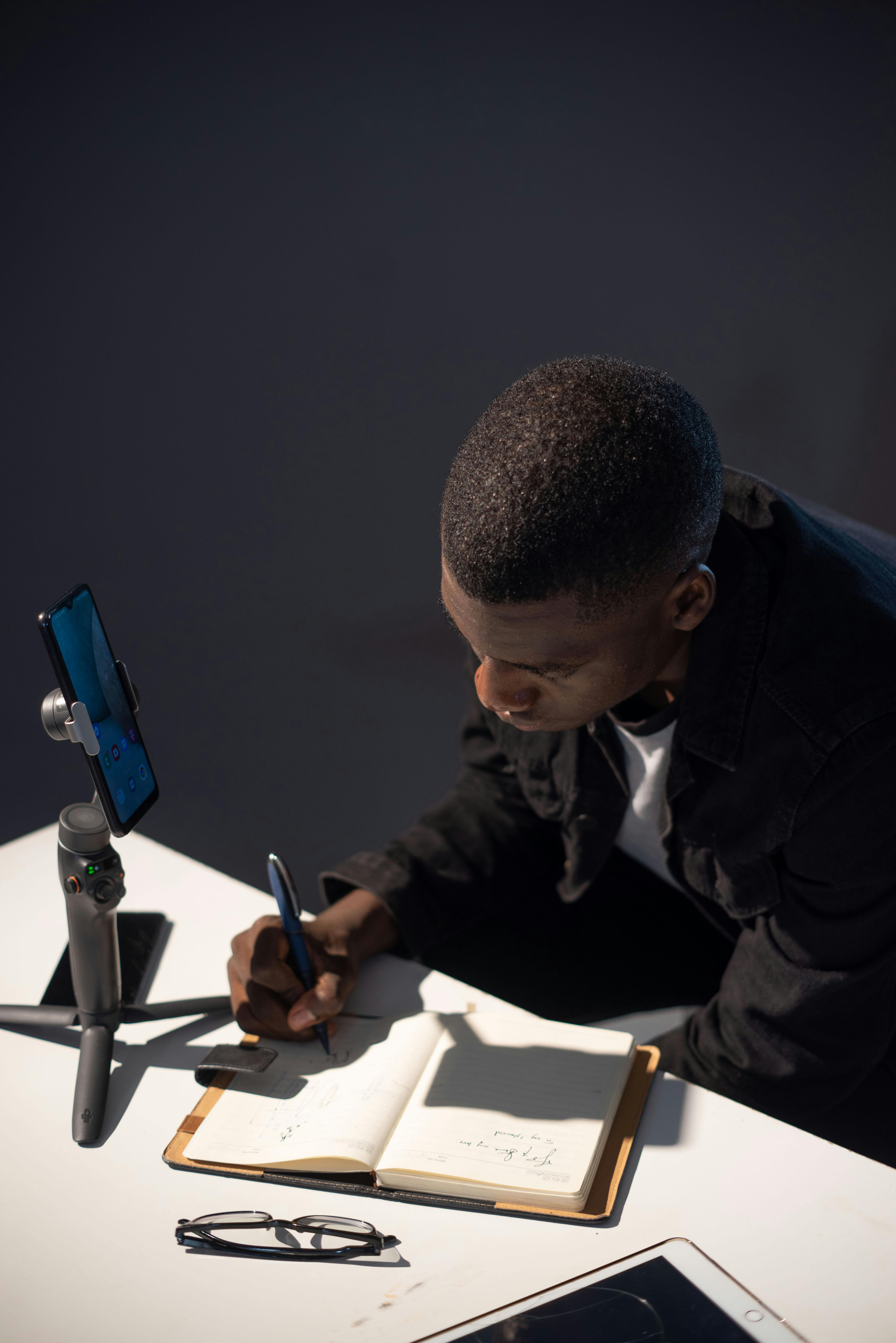 A man is writing on a tablet while sitting at a desk and looking at his phone