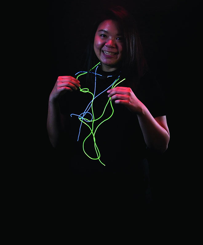 A person with long dark hair holds a neon green neon light string in front of their chest. The string is formed by multiple neon light trails. The person is smiling and wearing a black shirt. The background is entirely black.