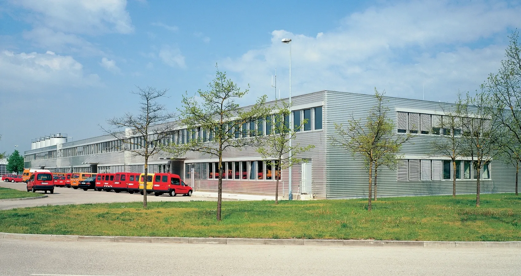 Moderne Lagerhalle mit parkenden roten und gelben Lieferwagen vor der Fassade unter blauem Himmel mit wenigen Wolken.