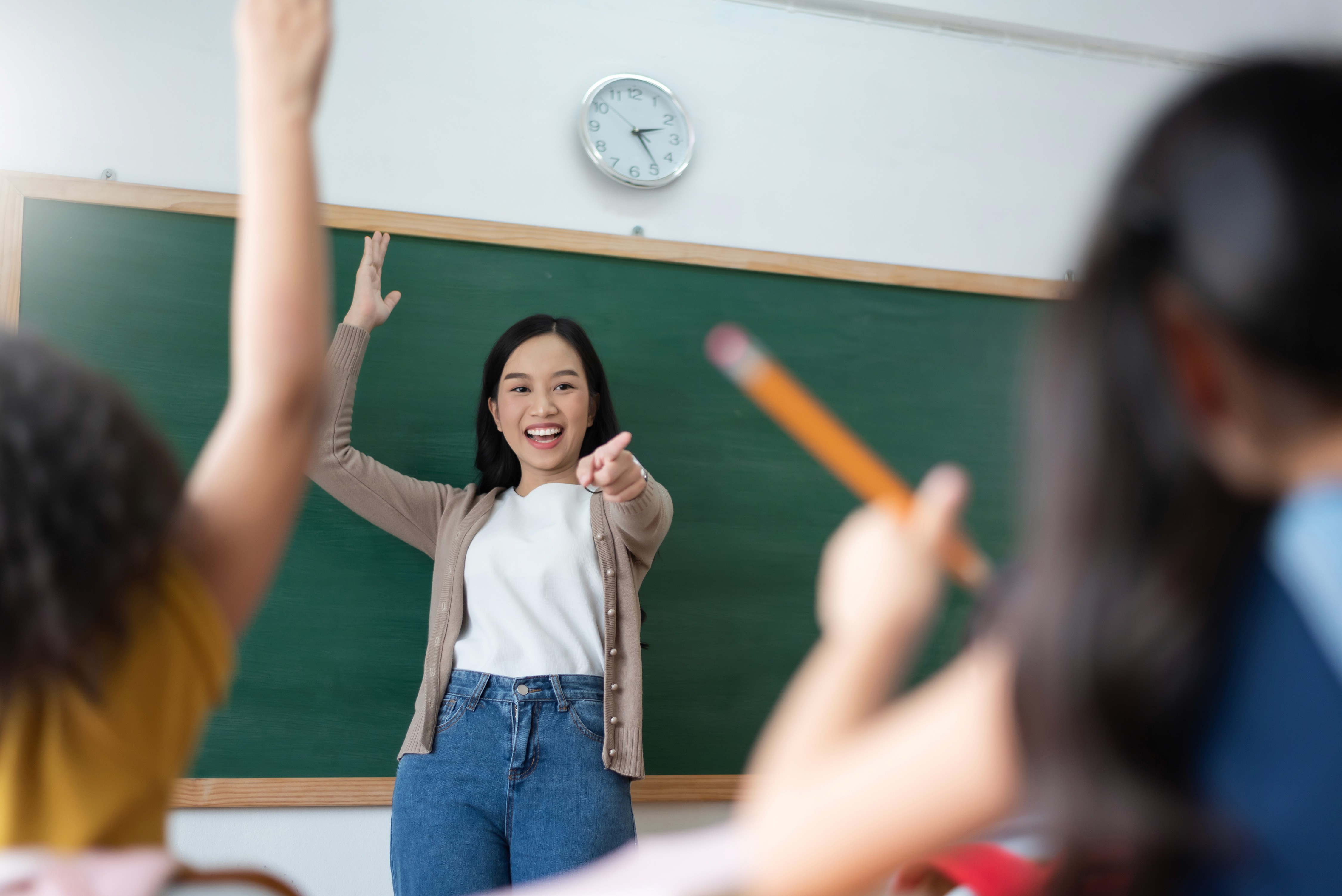 A smiling teacher stands in front of a green chalkboard with a clock above, engaging with students who are eagerly raising their hands, one holding a pencil.