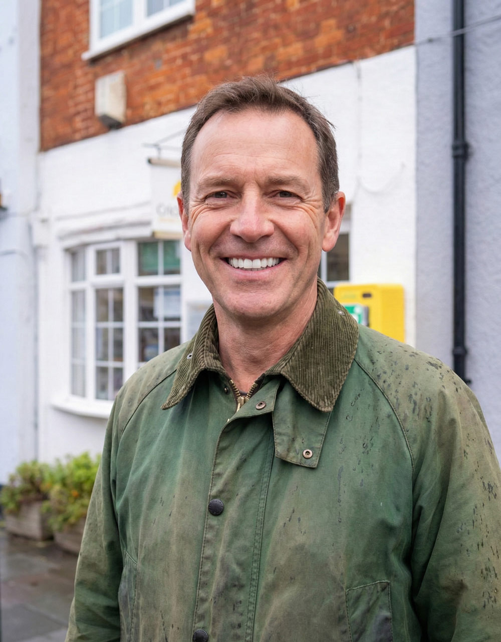 A smiling male patient in a green wax jacket stands outside the brick and white building of Cricklade Dental Practice.