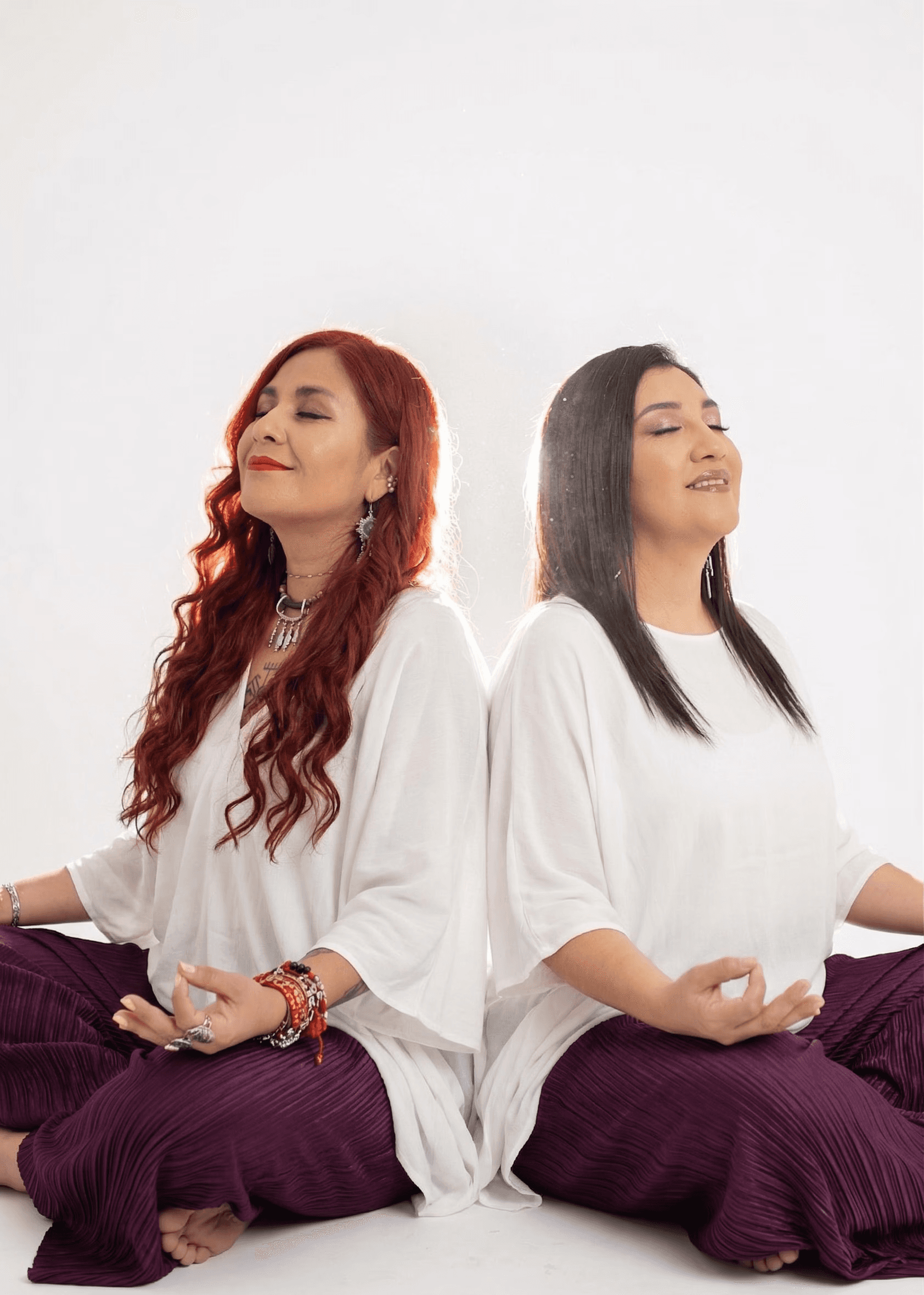 A circle of people sitting cross-legged, meditating with hands in a prayer position. Soft, warm lighting.