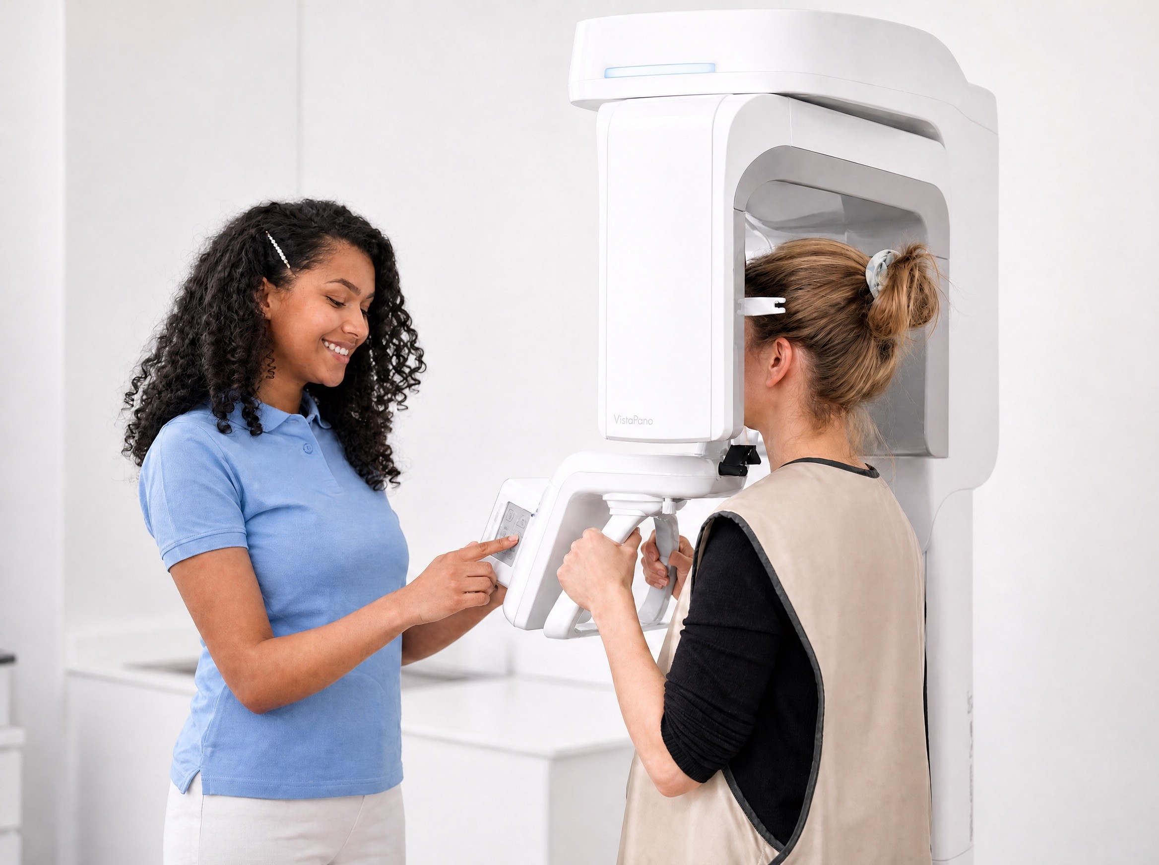 A female dental professional in a blue polo shirt adjusting a large, modern white panoramic X-ray machine for a female patient.
