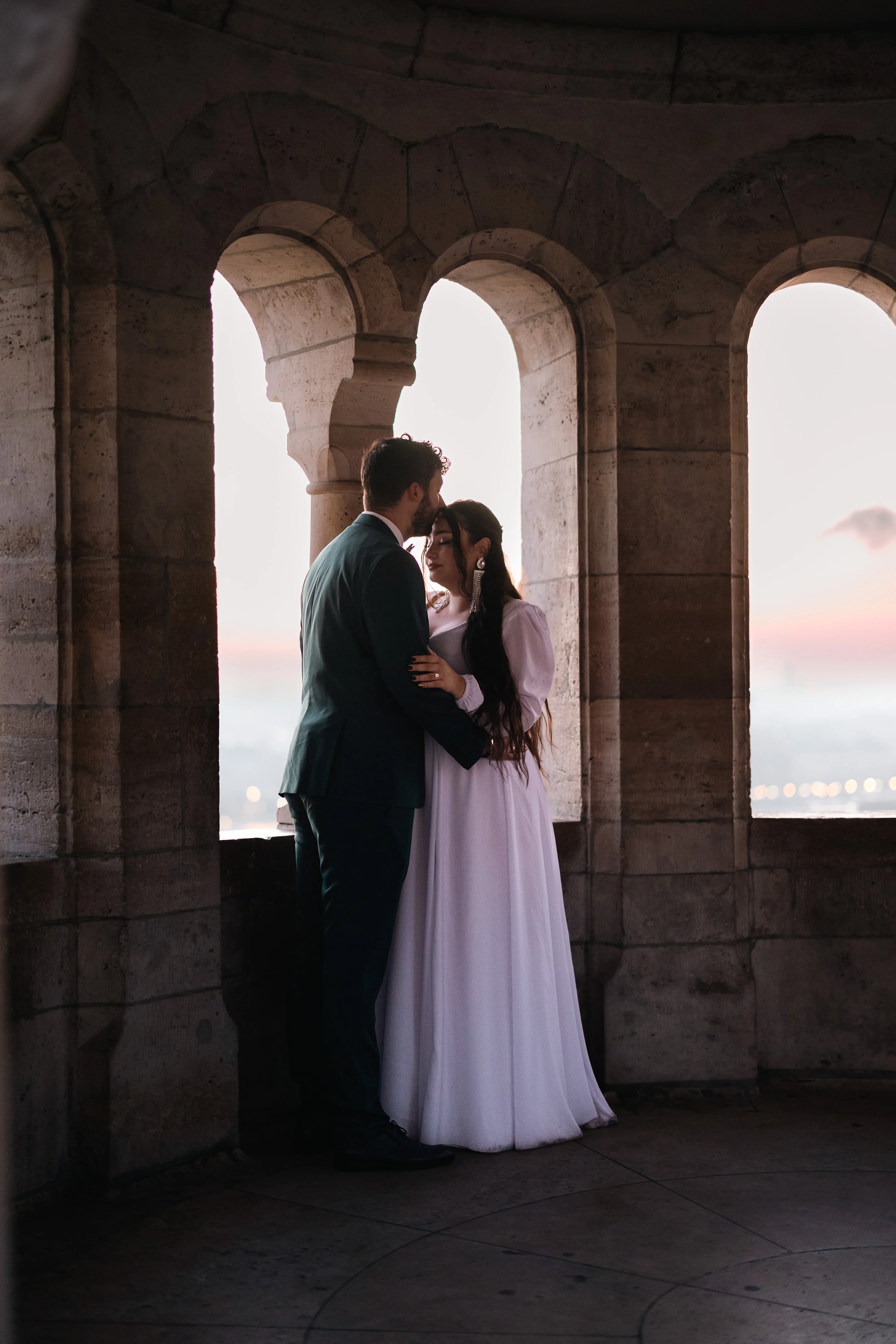 Menyasszony és vőlegény ölelkezve, romantikus kőablak mellett, naplementében. Budapest Halászbástyánál