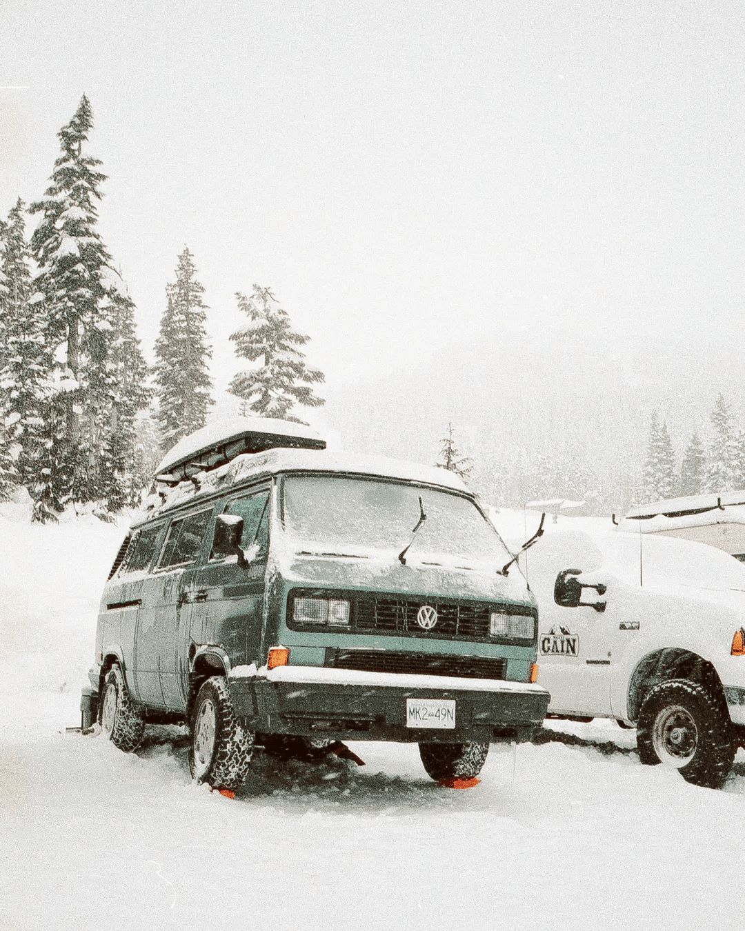 A snow-covered vintage VW van parked beside a pickup truck during a snowstorm