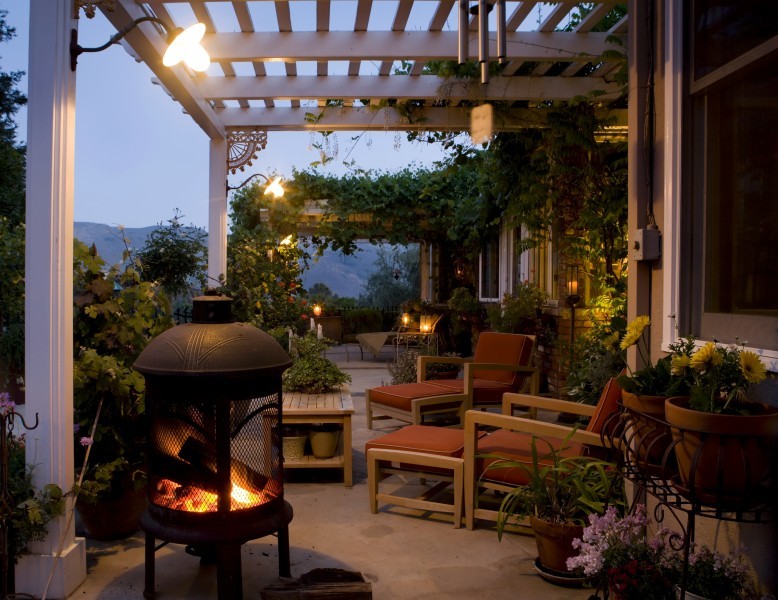 outdoor patio with an elegant wooden pergola, chairs, table, and fire pit surrounded by potted plants and flowers in vases