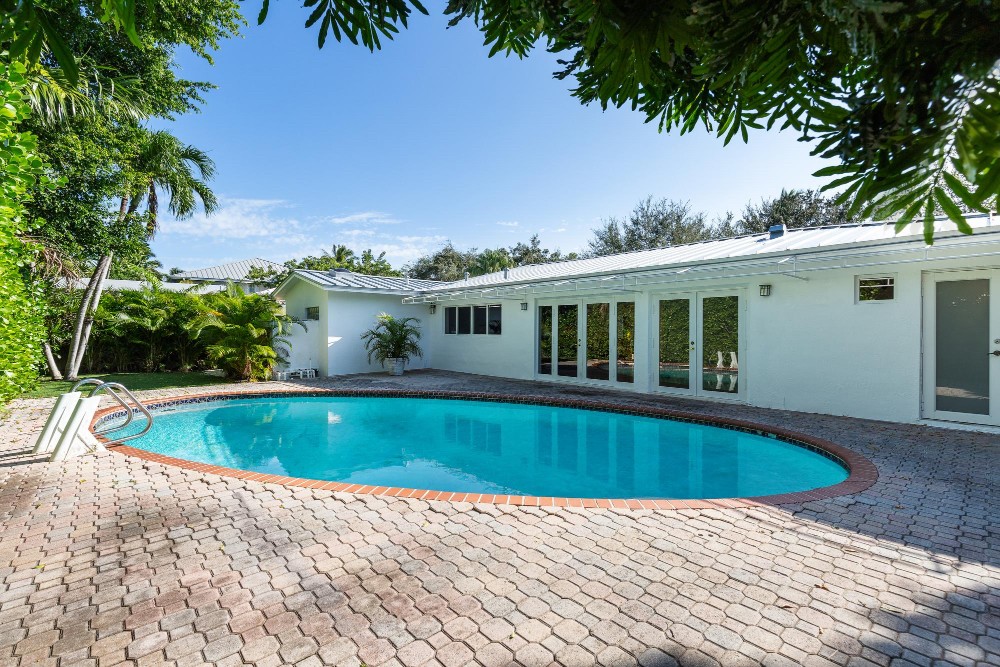 Residential backyard pool beside a white single-storey home with large windows and tropical landscaping.