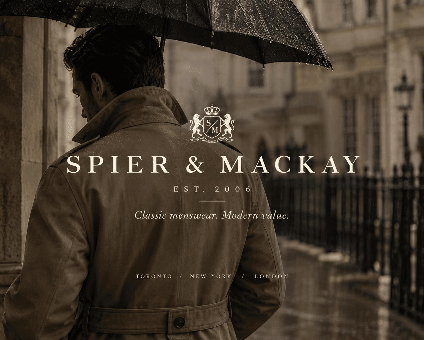 A back view profile of a man wearing a beige belt-cinched trench coat with an upturned collar, holding an open black umbrella on a rainy European city street. The background features blurred classical stone architecture, wet pavement, and a black wrought-iron railing. The center of the image displays white text with the crest logo for "SPIER & MACKAY EST. 2006", the tagline "Classic menswear. Modern value.", and the locations "TORONTO / NEW YORK / LONDON".