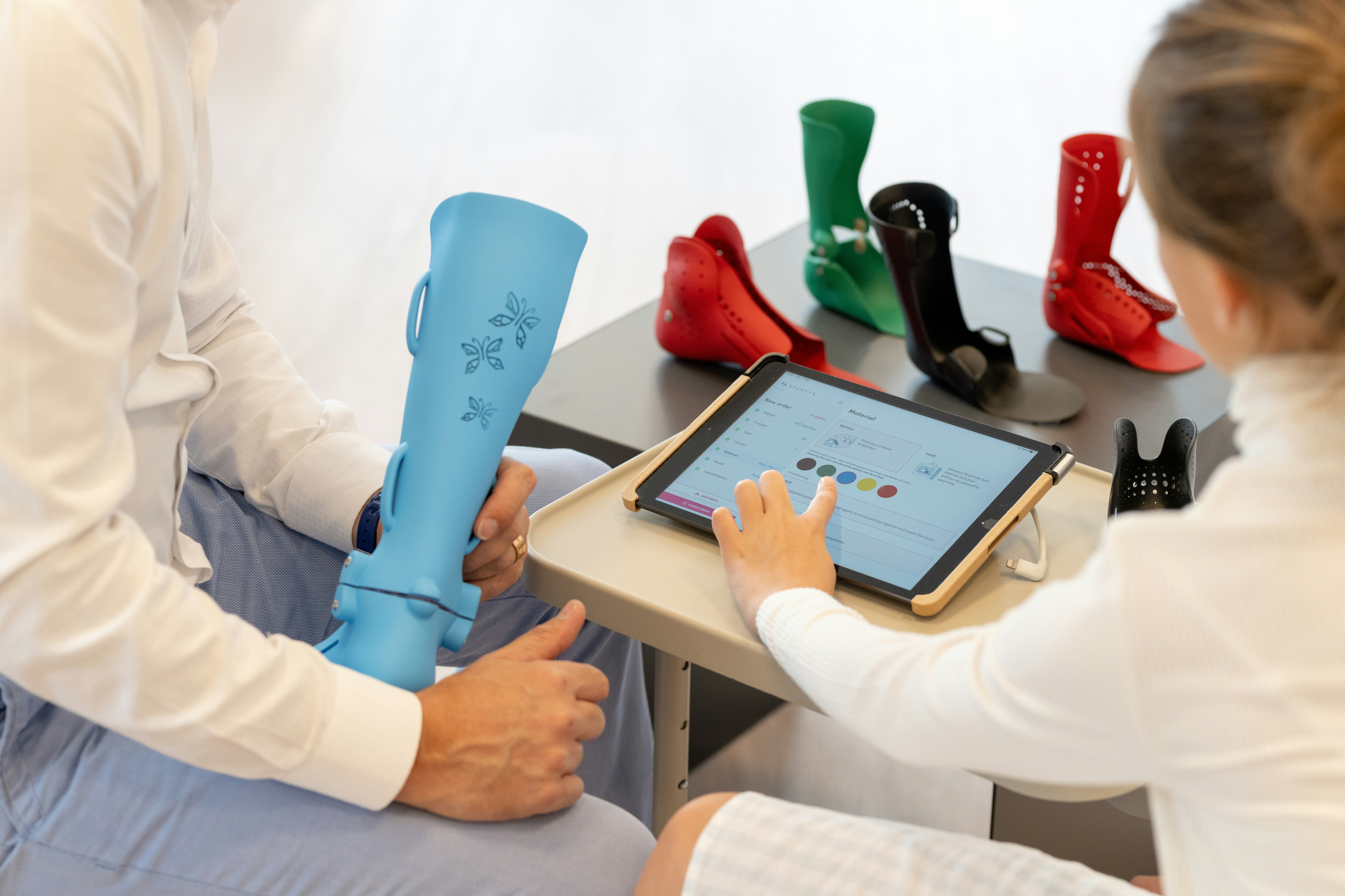 Two doctors discuss a blue orthotic brace while one holds a tablet displaying a color selection interface, surrounded by various colored braces on a table.