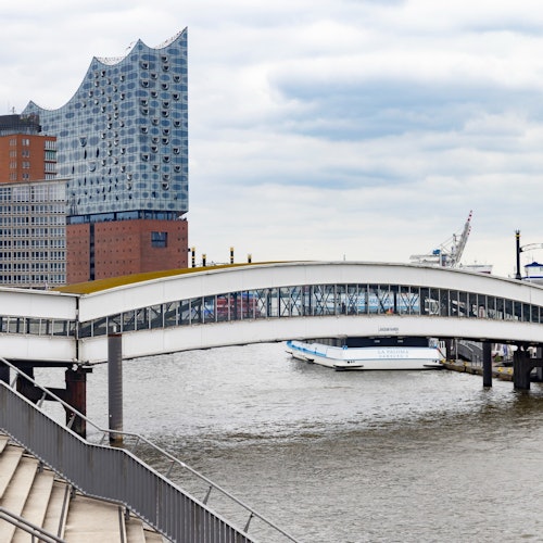 A modern building with a wavy roof, a pedestrian bridge over a river, stairs, and a docked boat under a cloudy sky.
